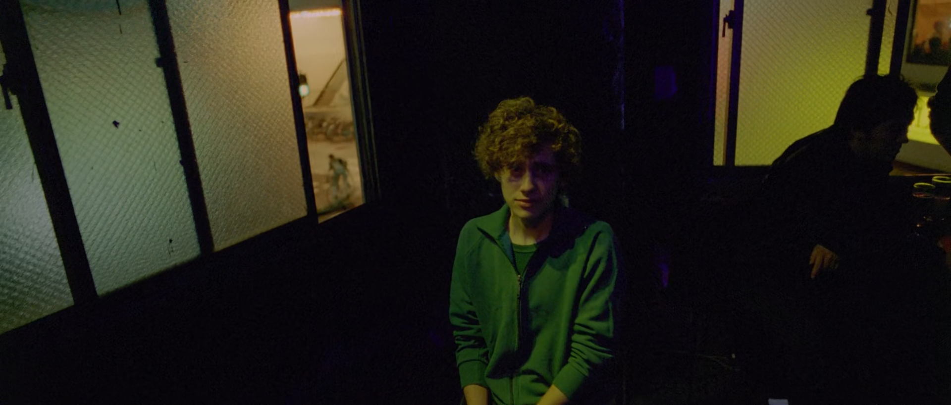 A young man wearing a green jacket sits in low light between pale textured glass panels, while another person appears as a dark silhouette on the right.