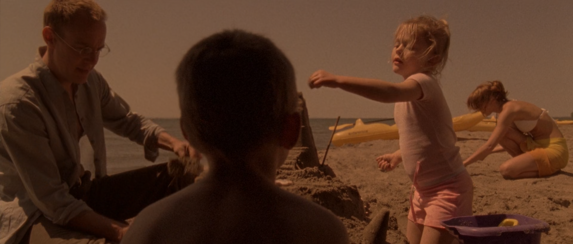 A family plays on a beach around a child seen from behind, with an adult man at left, a girl in pink at centre and two figures near the sand and water at right.