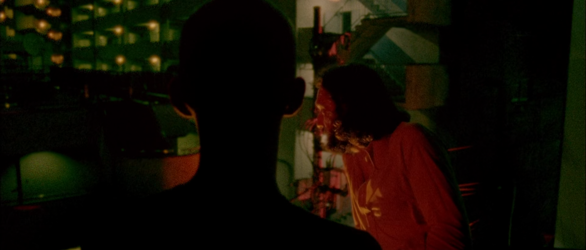 A dark head silhouette fills the centre foreground; behind it a long-haired man in red light stands at right and a green-lit room with small windows recedes at left.