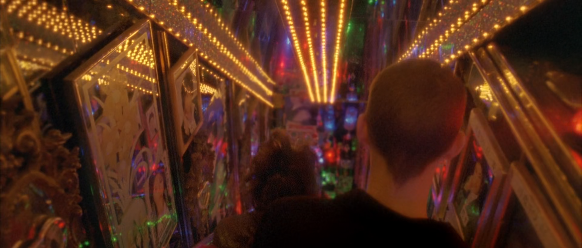Two people seen from behind descend a narrow staircase lined with mirrors, painted panels and converging rows of warm bulbs, with small coloured lights ahead.