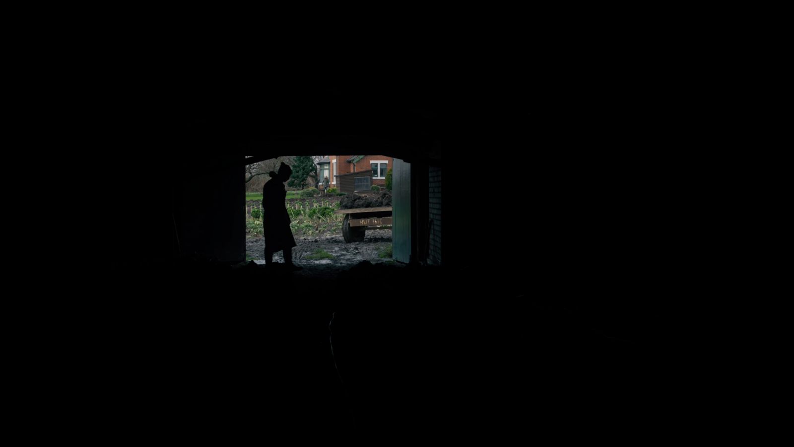 A small distant doorway and a standing silhouette remain visible inside an almost black shed.