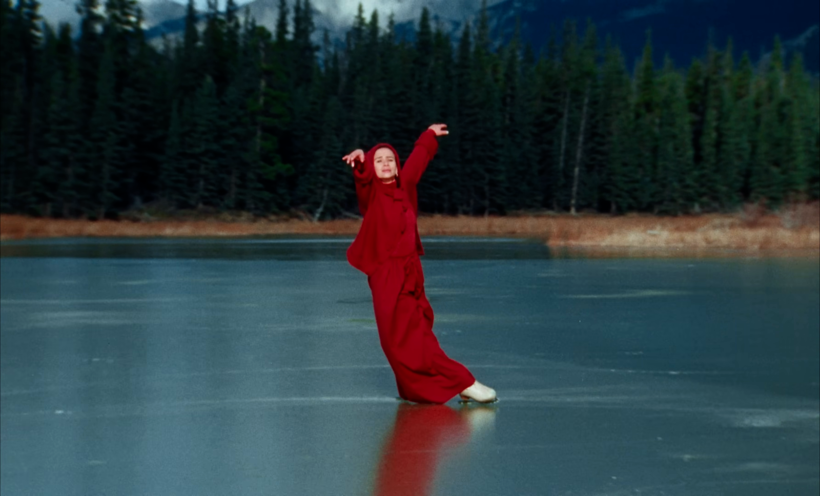 A skater in a red costume stands over a red reflection and fine marks on blue-green ice, separated from a dark forest.