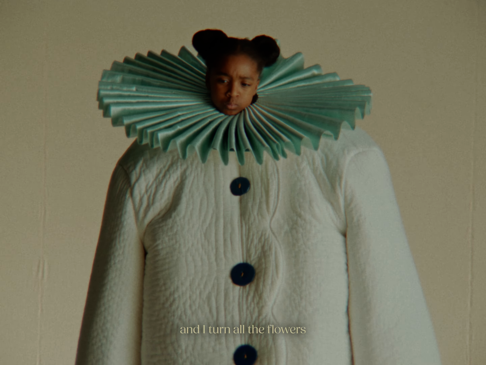 A child wears a pale textured costume and blue-green pleated collar against a warm cream backdrop.