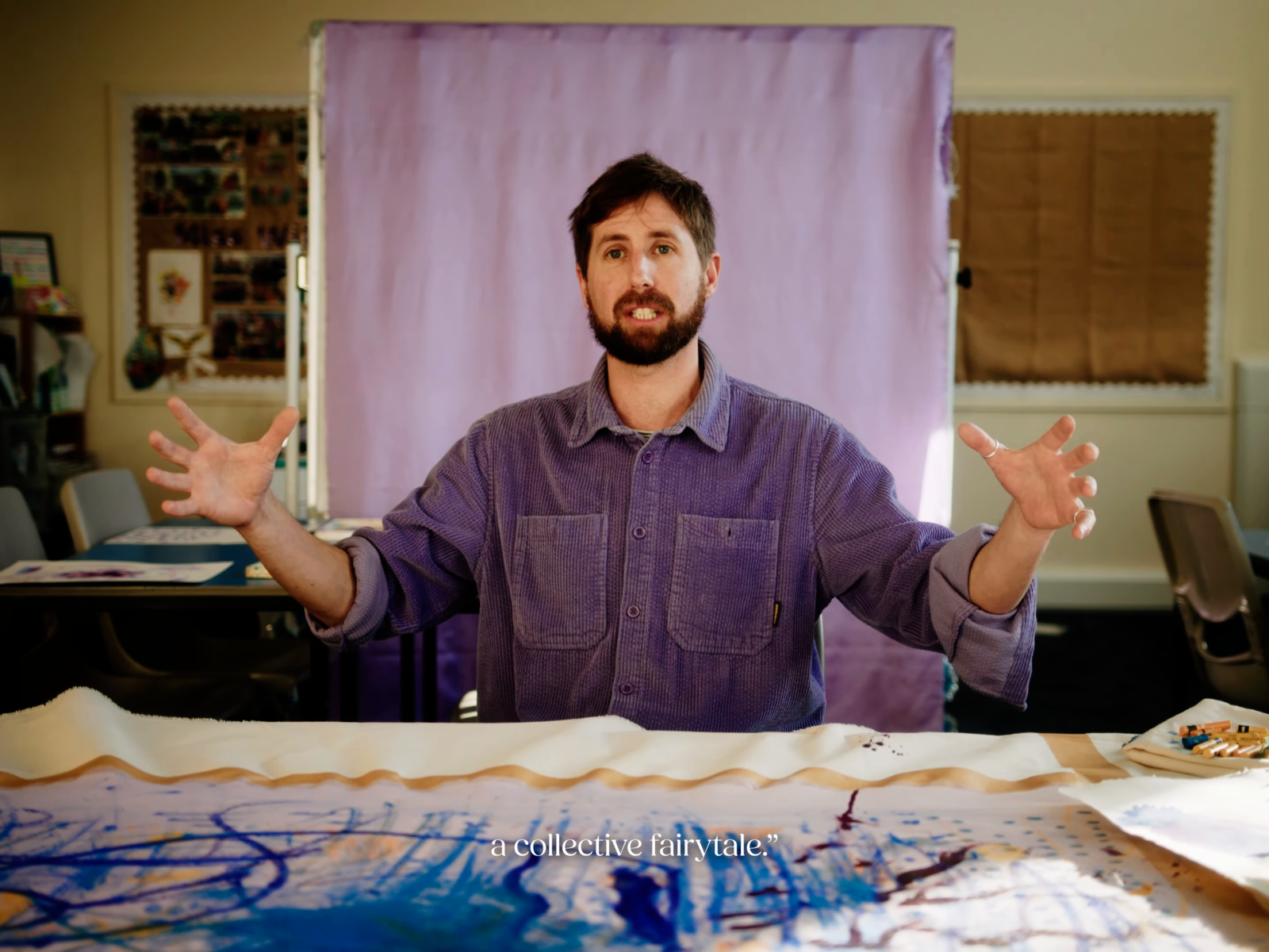 A man in a violet shirt sits behind painted fabric in front of a pale violet cloth backdrop; small white subtitles overlay the fabric below.