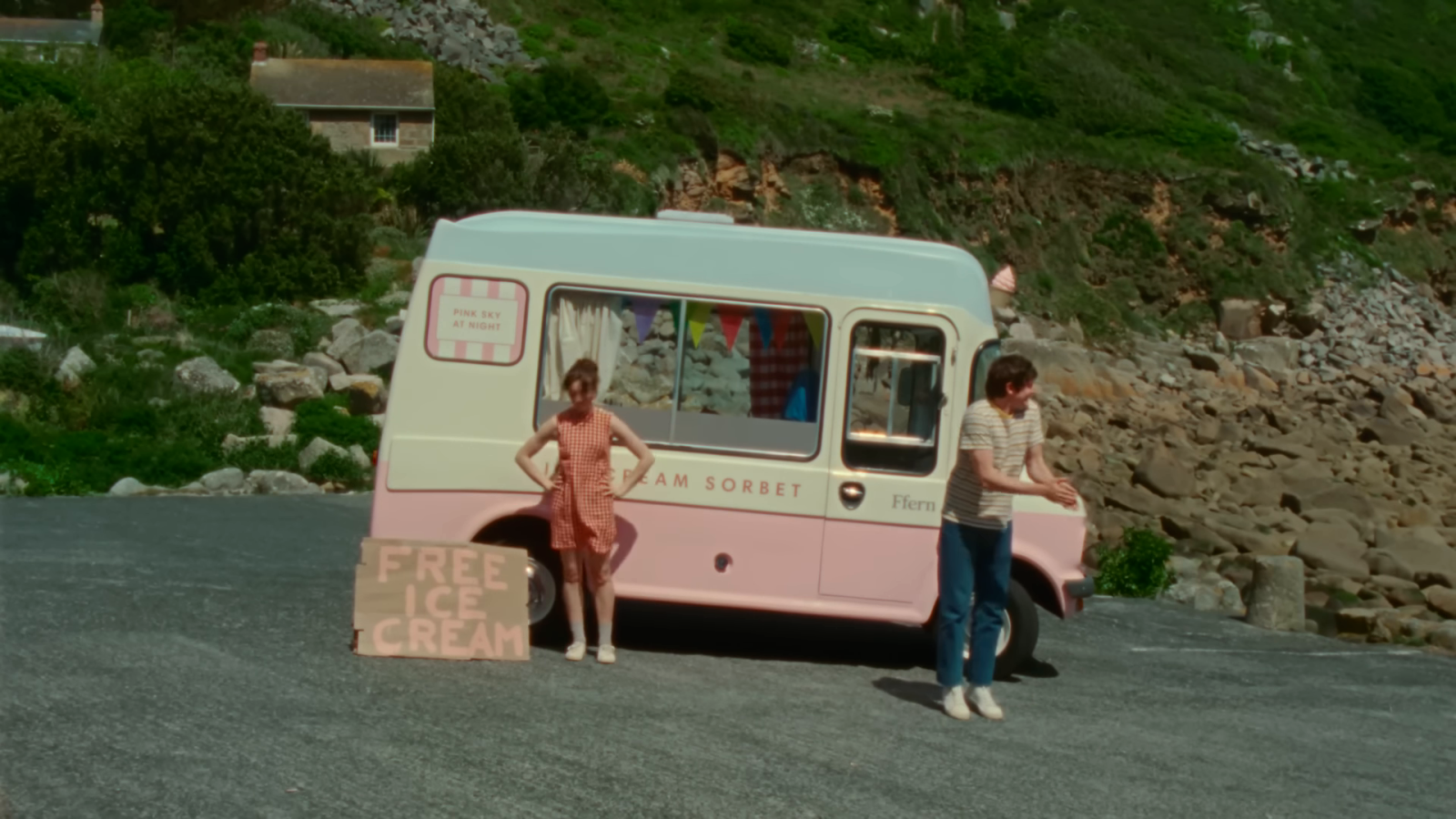 Two figures and a sign stand beside a pink-and-cream van against a rocky green hillside.