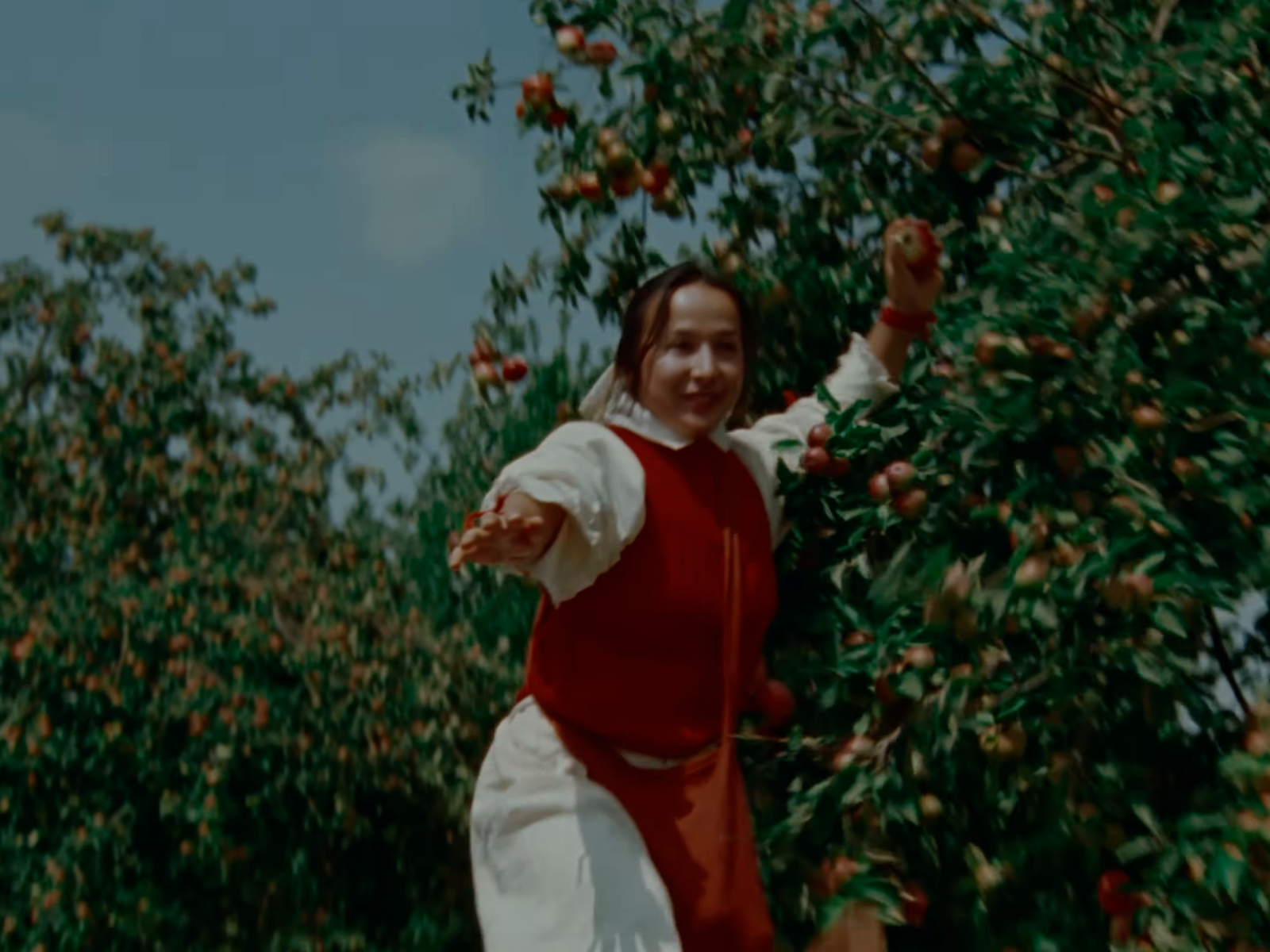 A figure in a red waistcoat and cream sleeves extends an arm against green apple trees.