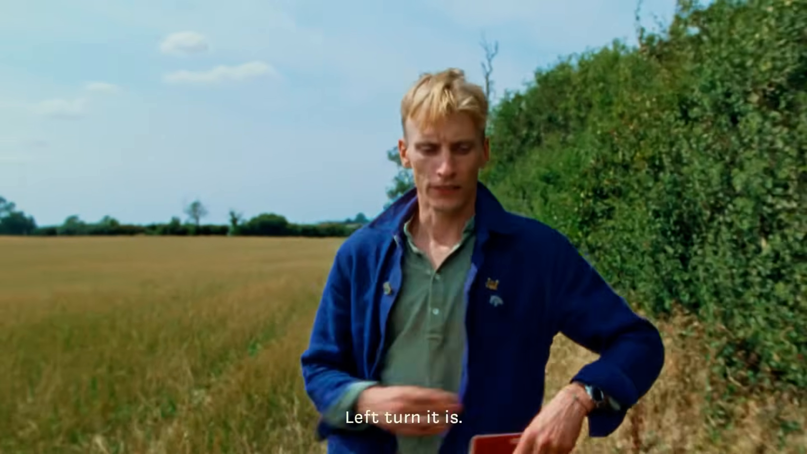 A man in a dark blue coat and green shirt stands in a gold field beneath blue sky.
