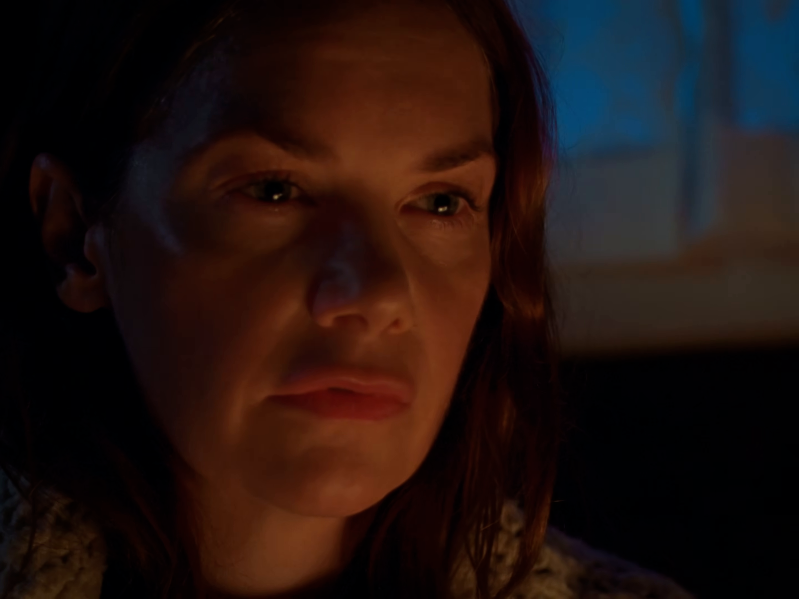 An orange-lit face is set against a deep blue room with much of the frame near black.