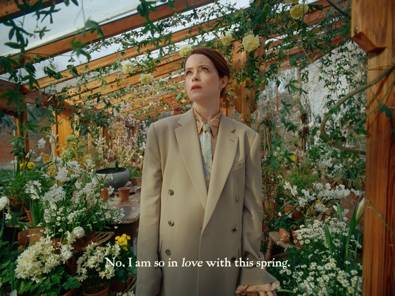 A woman in a beige suit stands among greenhouse flowers and timber bars, with a light serif subtitle across the lower centre.