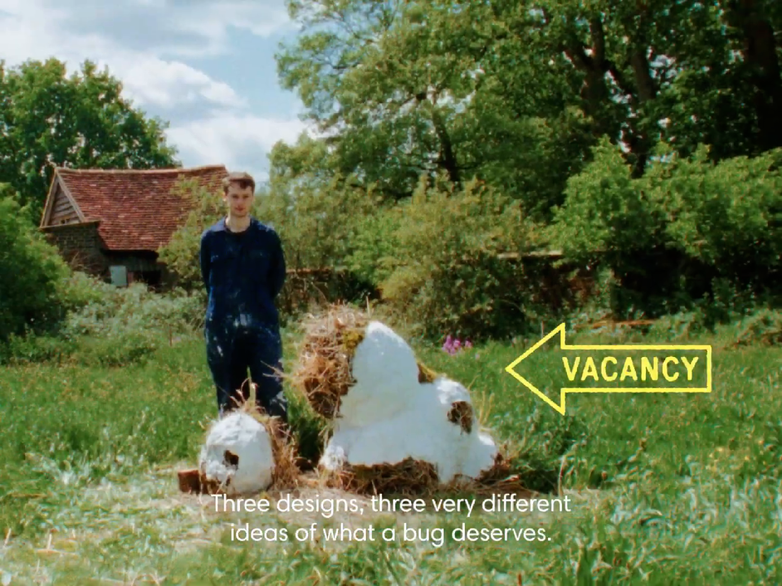 A designer in blue stands behind a pale insect-hotel structure in a meadow, with a yellow vacancy arrow at the right and white subtitles below.