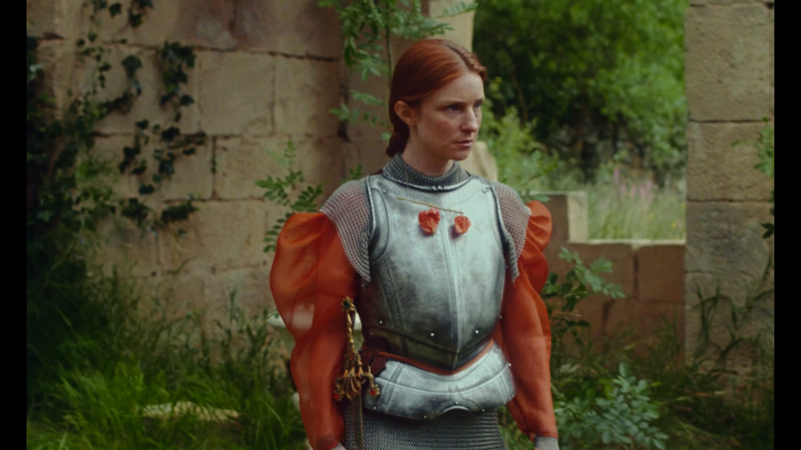 A red-haired figure wears silver armour over orange sleeves against pale stone and green vegetation.
