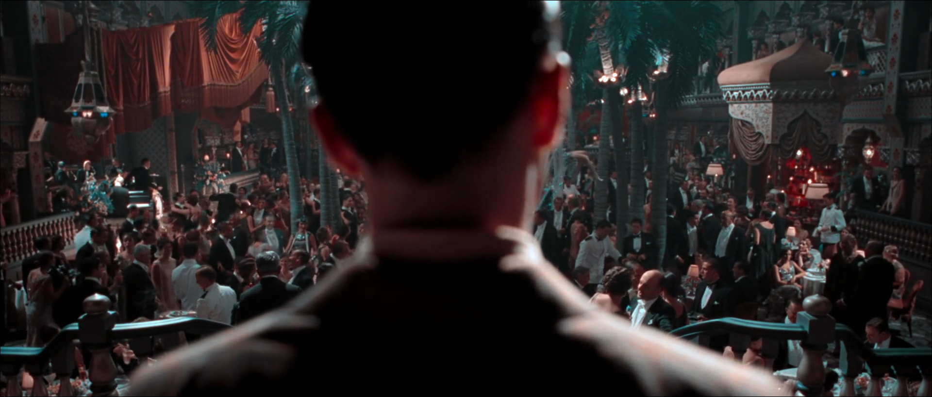 Seen from behind a dark-haired man at a balcony, a packed nightclub floor of men in tuxedos and women in evening dress among palm trees, with red drapes at upper left, a domed canopy at right and small lamps glowing across the room.