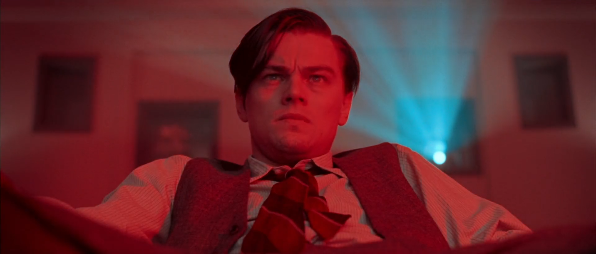 A young man in a striped shirt, dark waistcoat and loosened tie looks up and past the camera in a room bathed in red light, with dark picture frames on the walls and a pale cyan projector beam shining toward him from behind at right.