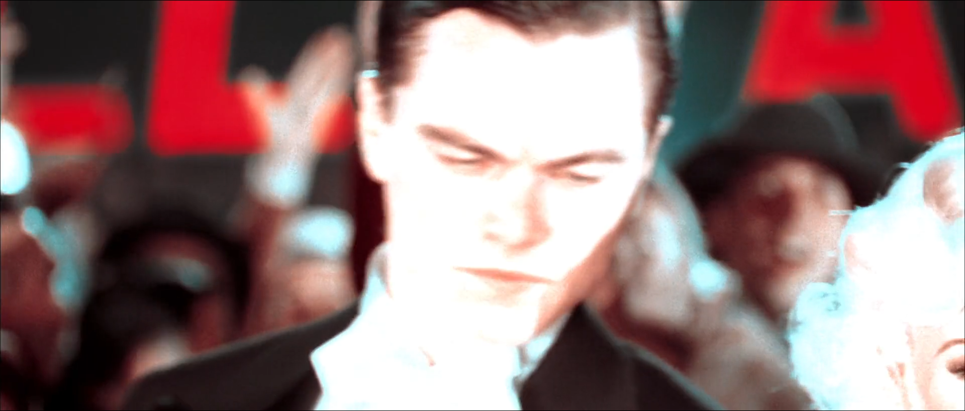 A young man in a dark suit and white shirt looks down in close-up, his face overexposed nearly to white, in front of blurred red capital letters and a crowd, with a woman's platinum hair at the right edge of the frame.