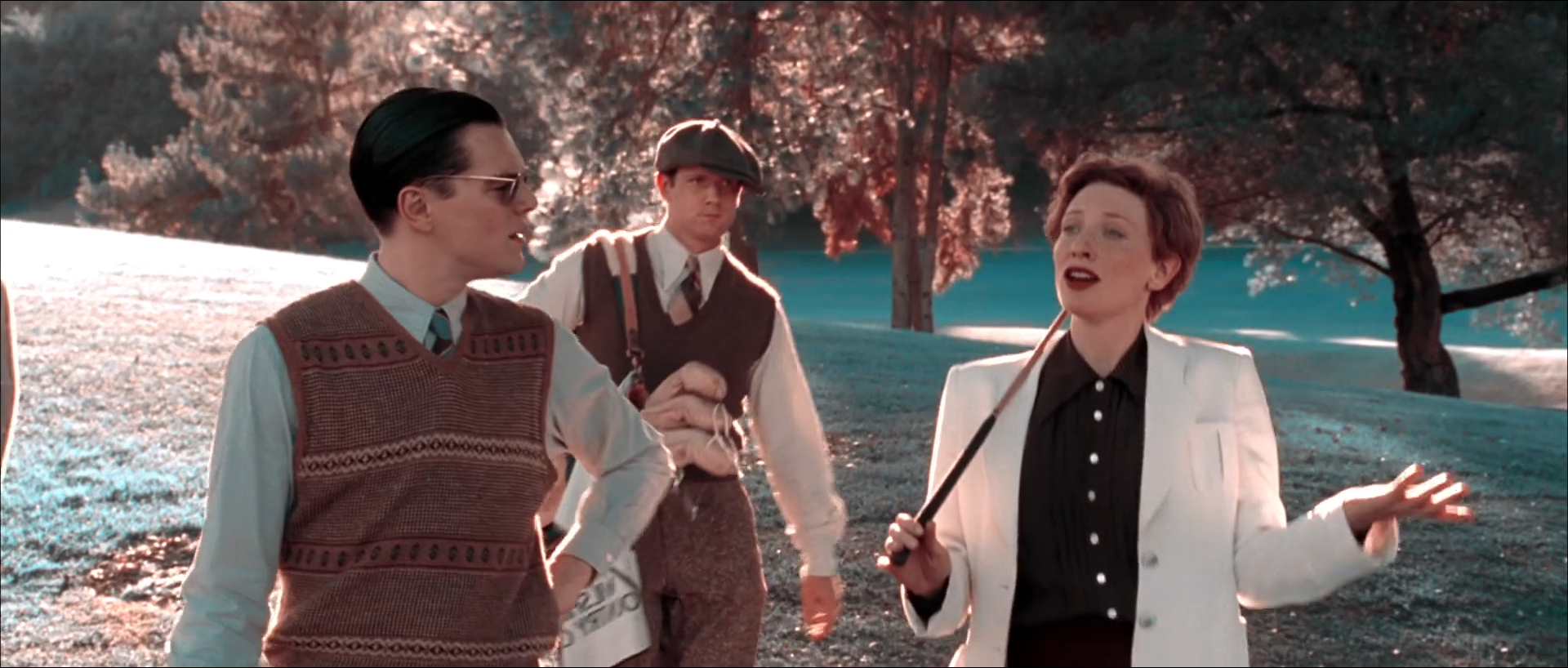 Three people walk on a sunny golf course: a man in sunglasses and a patterned sweater vest, a young caddy in a flat cap with a bag, and a red-haired woman in a white jacket and dark blouse gesturing with a golf club, in front of pink-leaved trees and pale cyan grass.