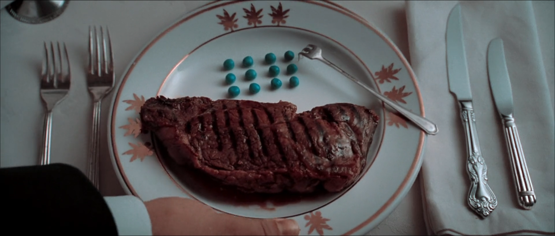 Top-down view of a white plate with a copper-coloured leaf border holding a grilled steak and twelve round peas in rows, with silver forks at left, knives and a white napkin at right, and a hand in a black sleeve at the lower left.