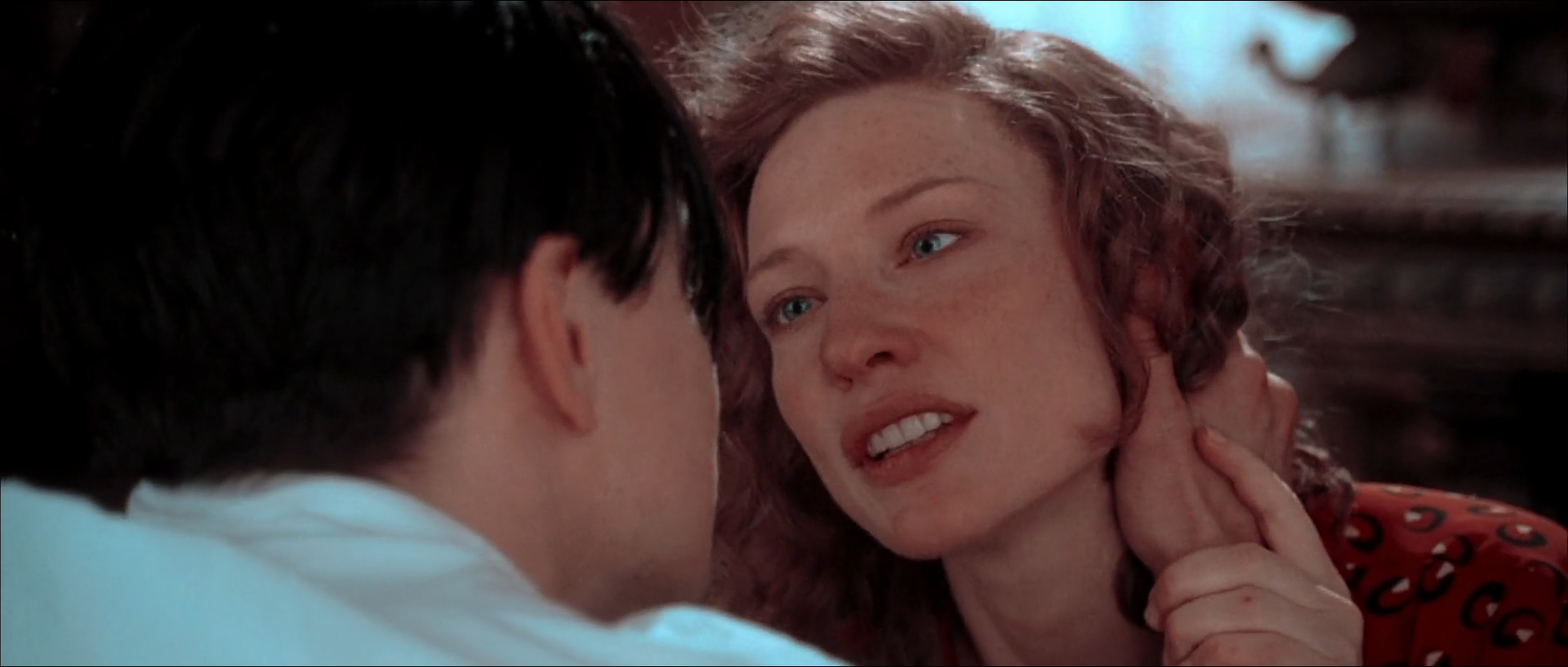 Close-up of a red-haired, freckled woman with blue-grey eyes looking up and speaking to a dark-haired man seen from behind at left, her hand at her neck, in a red patterned dress, with a soft cool window light behind her.