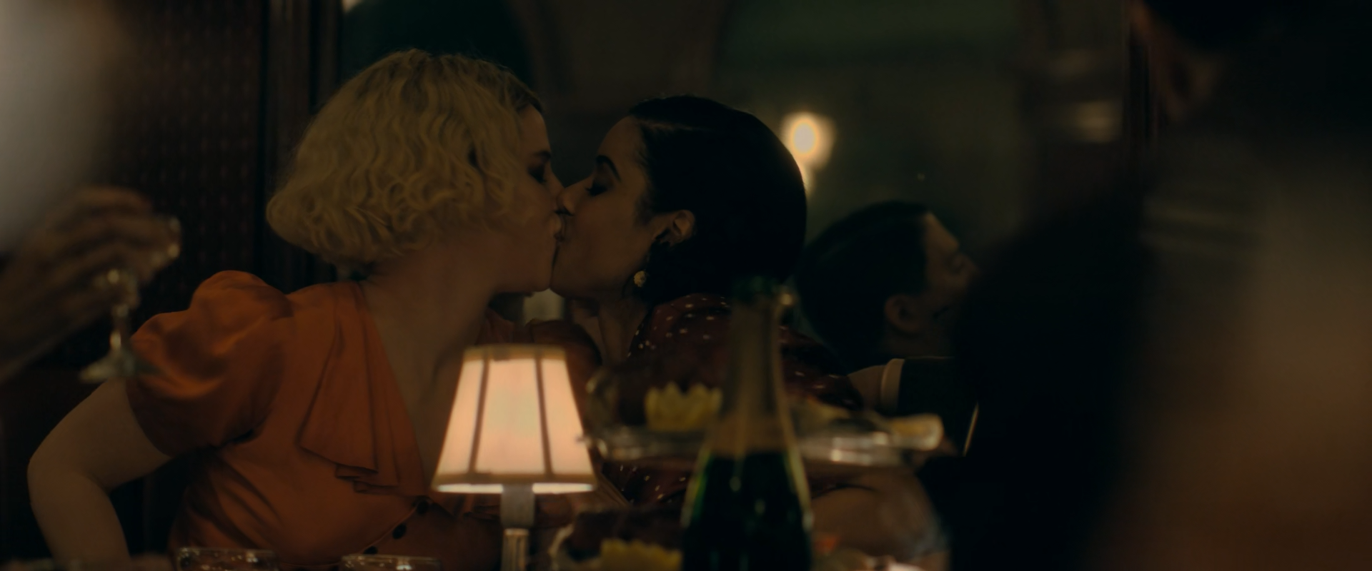 Two women kiss across a dim restaurant table: one blonde with curled hair in an orange satin blouse, the other dark-haired in a dotted dress, beside a small white-shaded table lamp and a green bottle, with other diners blurred in the dark.