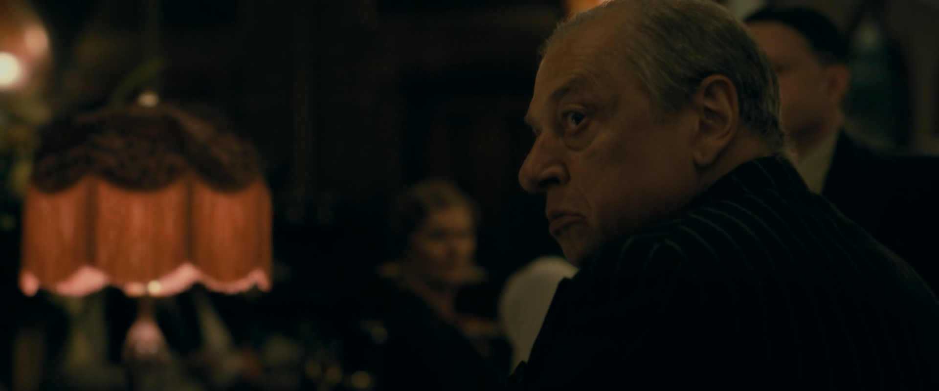 An older grey-haired man in a dark pinstriped suit looks back over his shoulder in a dark room, with a glowing rose-orange fringed lampshade and a bright bulb out of focus at left and two blurred people behind him.