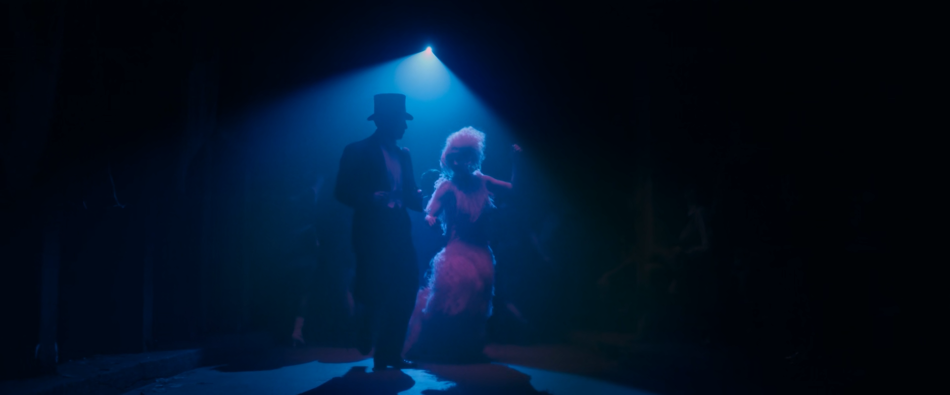 A man in a top hat and tails and a woman in a feathered red dress dance in a cone of blue light falling from a single spotlight above, in an otherwise black nightclub with faint figures at the edges.