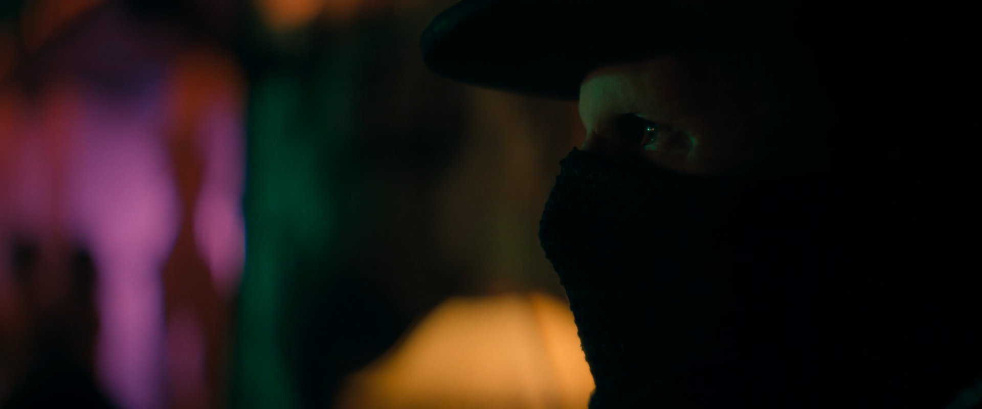 Close on a man in a black hat and black knitted face mask, only his eye and part of his brow lit, against blurred magenta, violet, green and amber lights at the left of the frame.