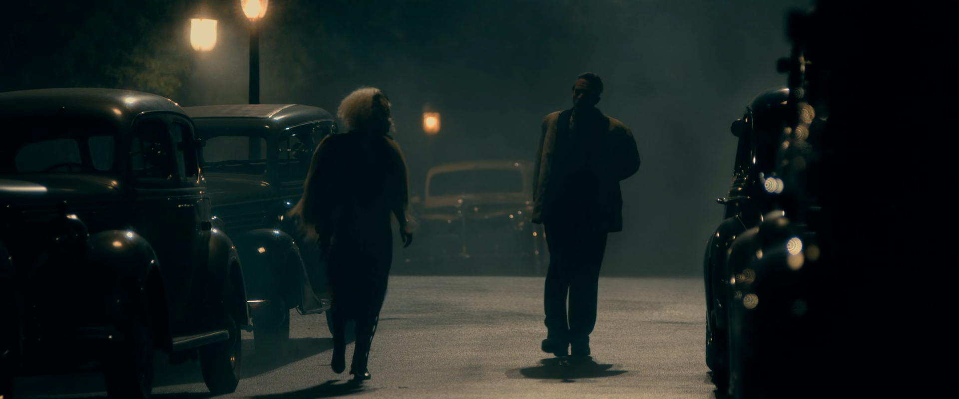 Two figures, a woman with pale curly hair and a man in a jacket, walk down a hazy night street between parked vintage cars, backlit by glowing streetlamps with another car in the fog at the far end.