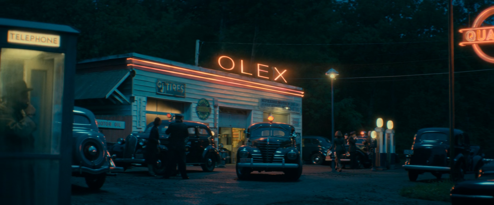 A roadside gas station at dusk with an orange OLEX neon sign, several dark 1930s police cars with red roof lights, uniformed officers, a man inside a lit telephone booth at left, and glowing globe fuel pumps at right in front of dark trees.