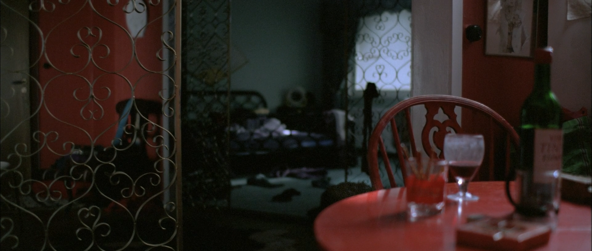An interior with a red round table, red chair, wine glass and bottles stands before ornate metalwork and a darker green-blue room beyond.
