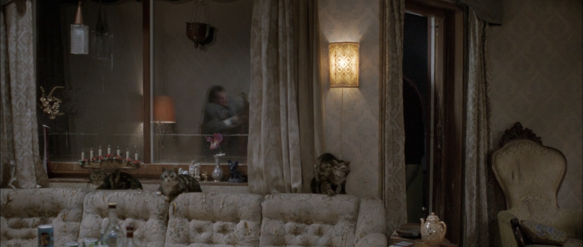 Several cats occupy a pale sofa and chair in an ornate cream room, with curtains, a lit wall sconce and blurred people visible in a darker glass or reflected area behind them.