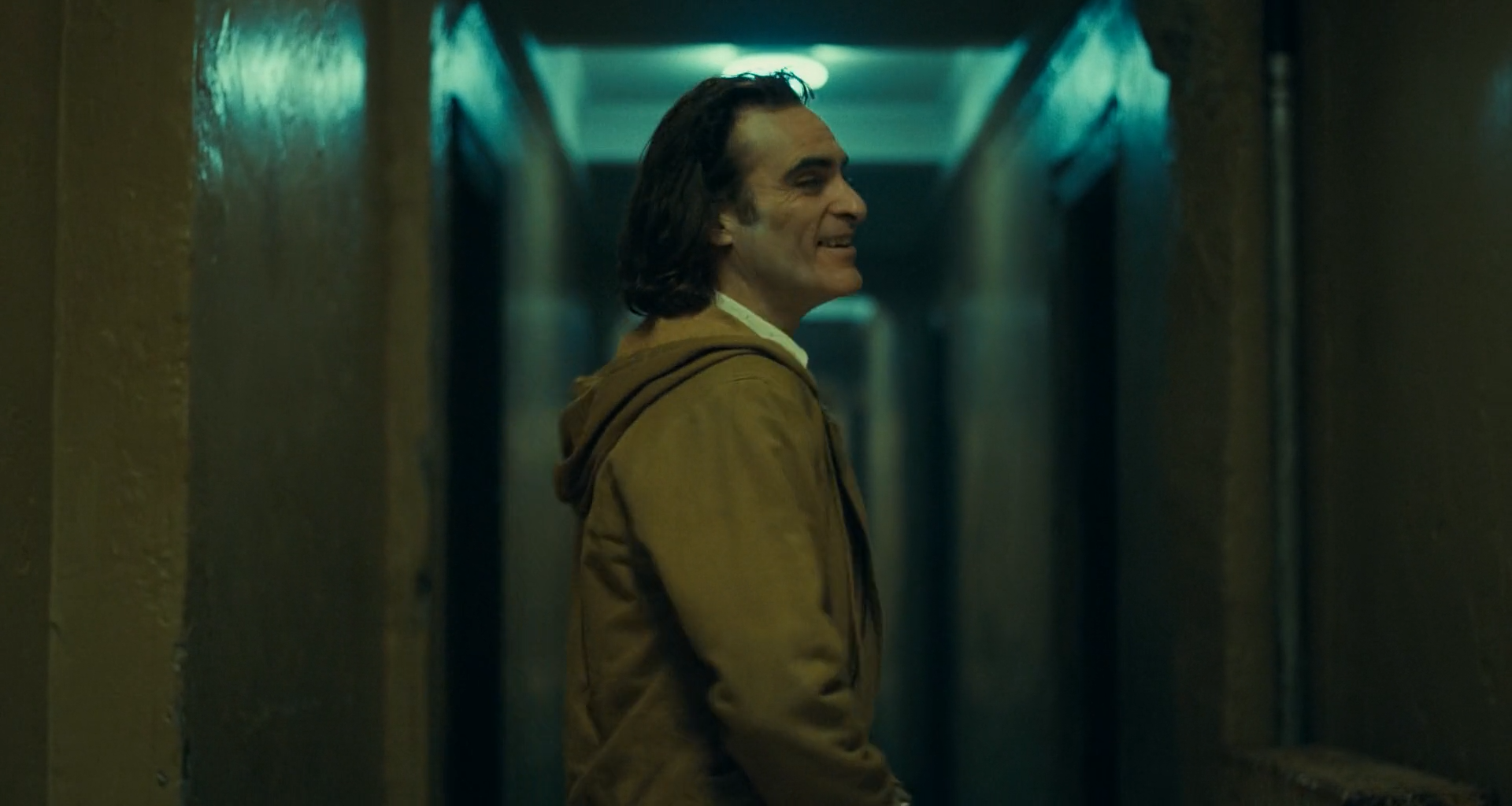 A man with shoulder-length dark hair in a brown hooded jacket stands in profile, smiling, in a narrow corridor with dark olive walls receding to a teal-lit ceiling fixture.