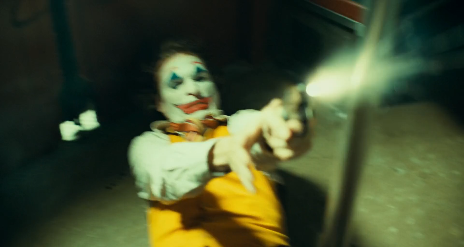 A man in white clown makeup with a red mouth and blue eye markings, a white shirt and mustard waistcoat, fires a revolver toward the camera inside a subway car, with a bright yellow-white muzzle flash, motion-blurred arm and a blurred pole in the foreground under green light.