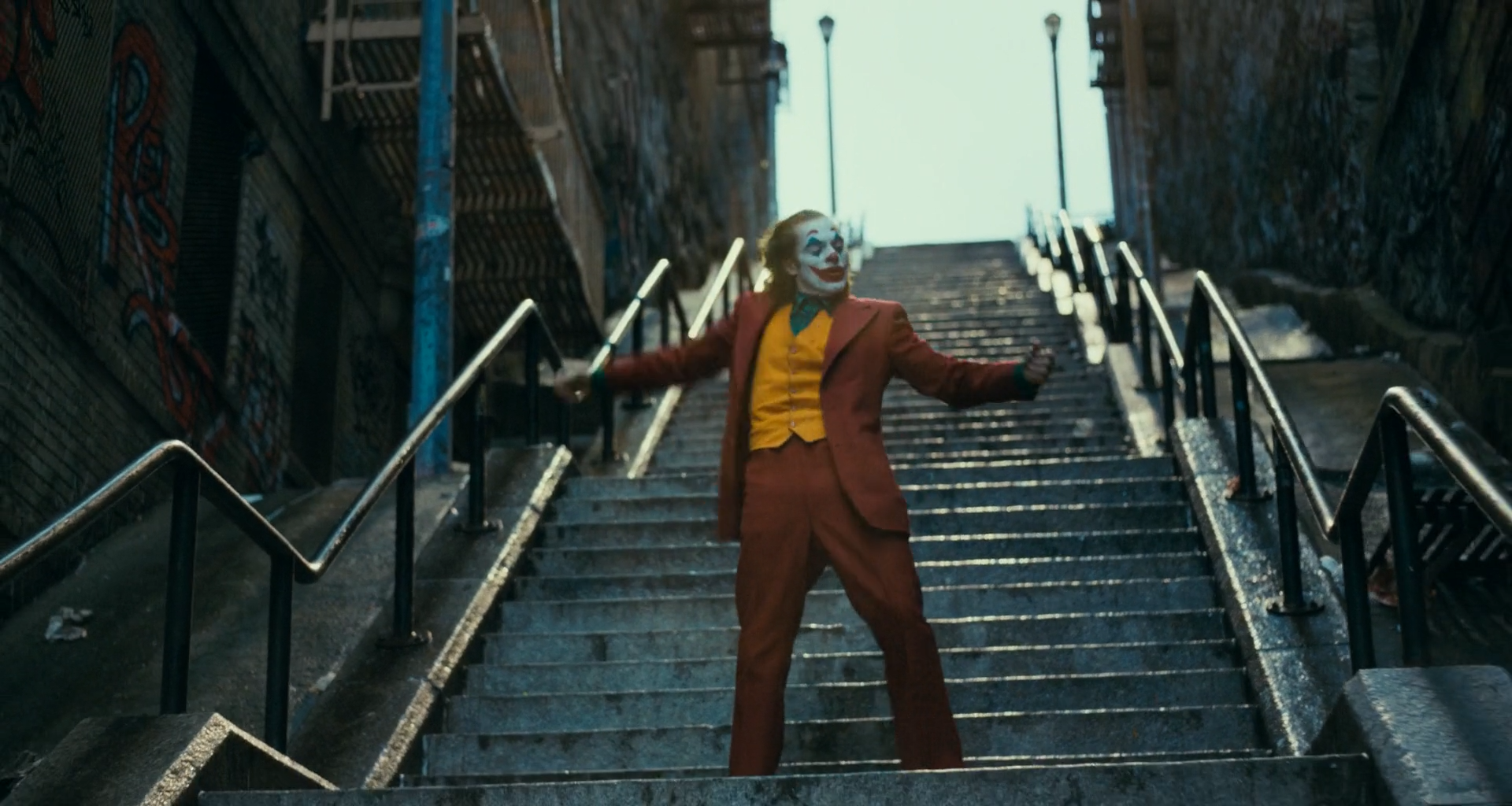 A man in white clown makeup, a rust-red suit, mustard waistcoat and green shirt dances on a long concrete outdoor stairway between graffiti-covered walls, with metal railings and two streetlamps at the top against a pale overcast sky.