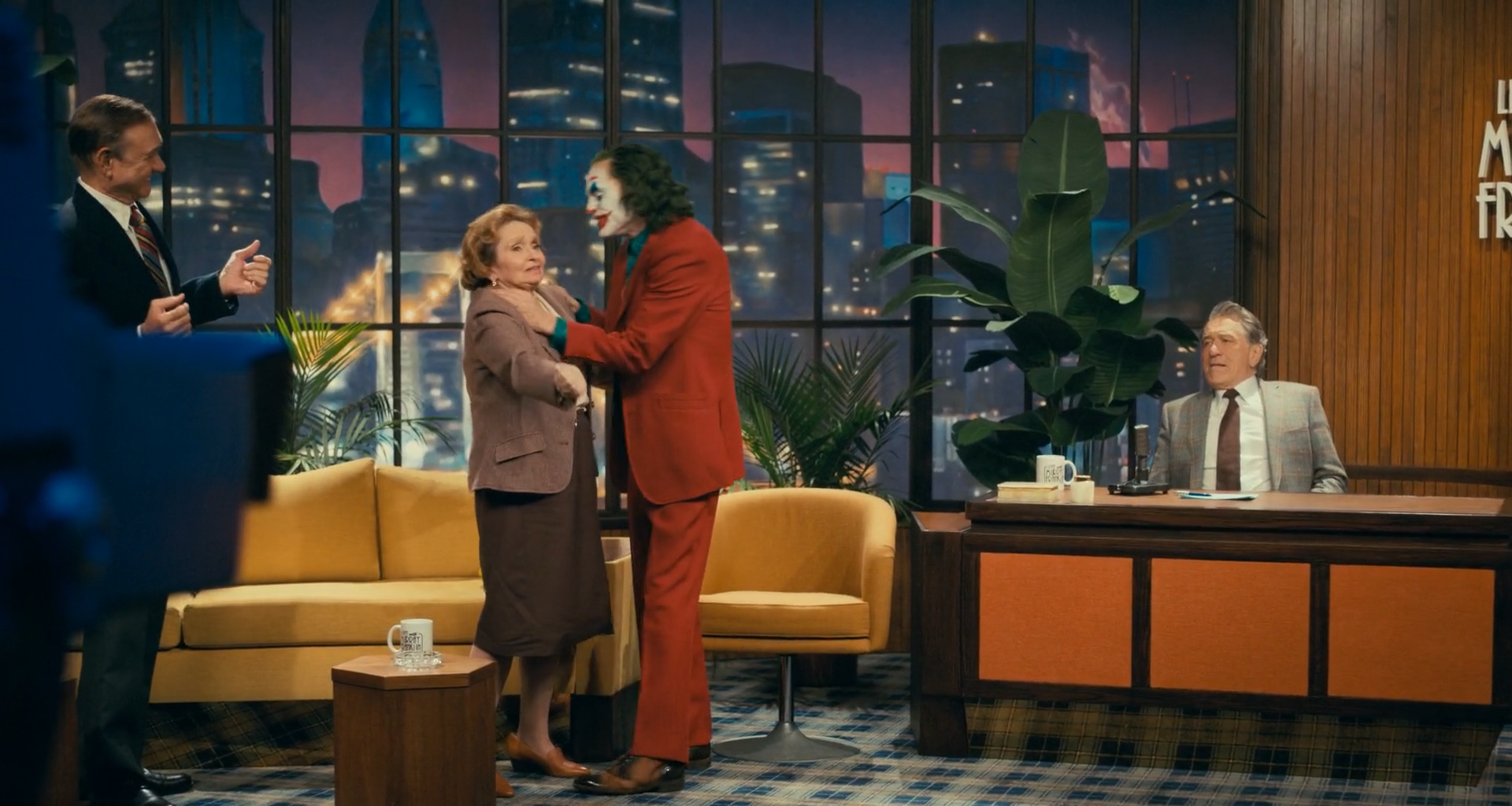 A 1970s television talk-show set with mustard sofas, a plaid carpet, potted plants, a wooden desk with orange panels where a host sits, a man in a rust suit and clown makeup holding an older woman by the shoulders, a man gesturing at left, a blue studio camera in the left foreground, and a painted night skyline with pink sky and lightning behind windows.