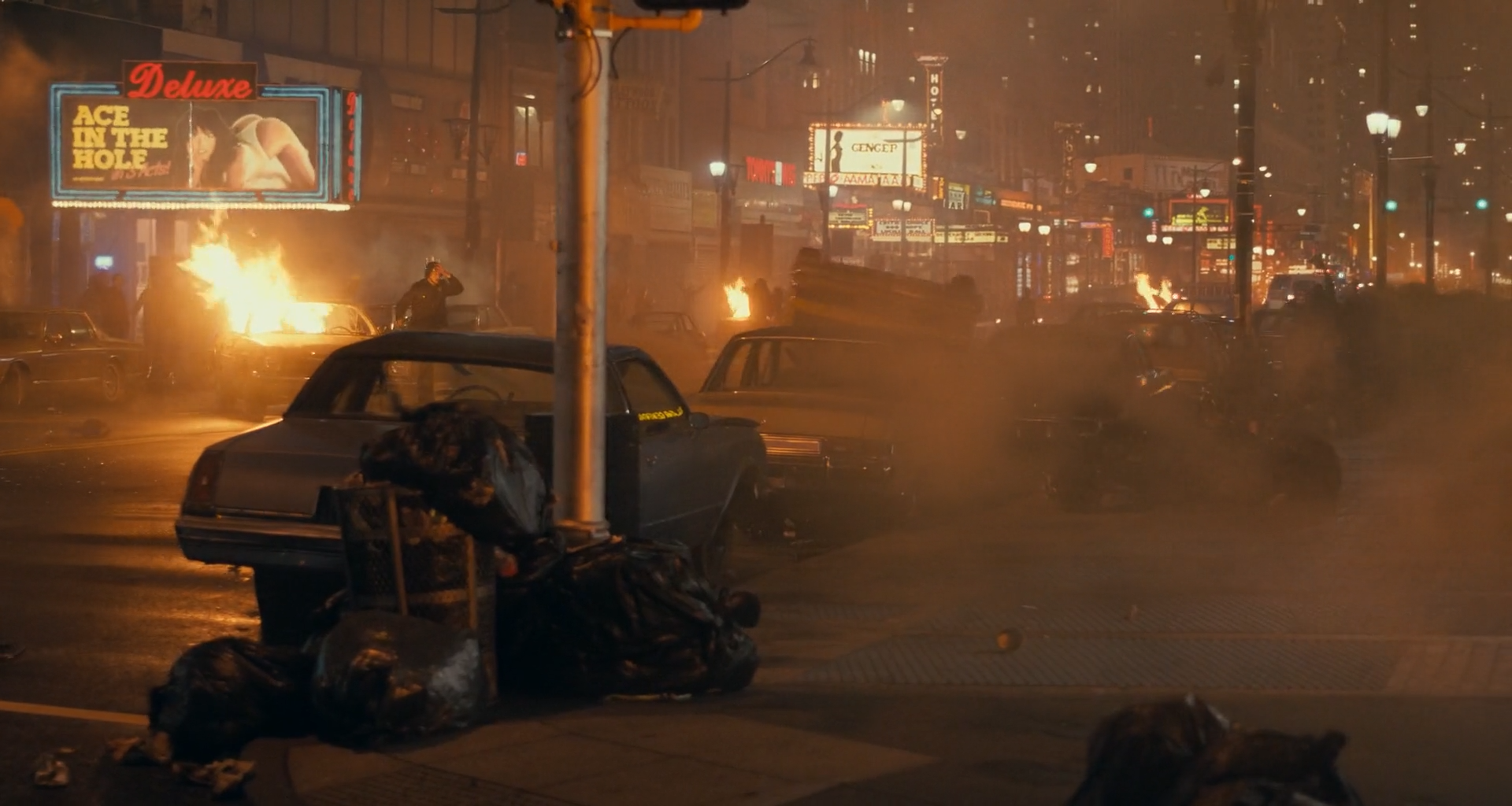 A smoke-filled city street at night with burning cars, rubbish bags piled around a lamp post in the foreground, a glowing billboard reading Deluxe ACE IN THE HOLE, a figure on a car, and streetlamps and neon signs receding into amber haze.