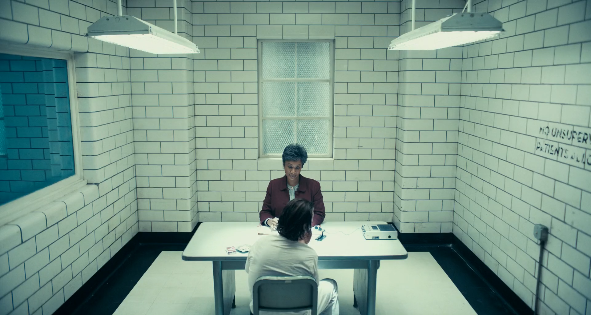 High-angle view of a white-tiled room with two hanging fluorescent lights, a frosted window on the back wall, a dark observation window at left and a stencilled notice at right, a woman in a maroon jacket sitting at a table facing a man in white seen from behind.