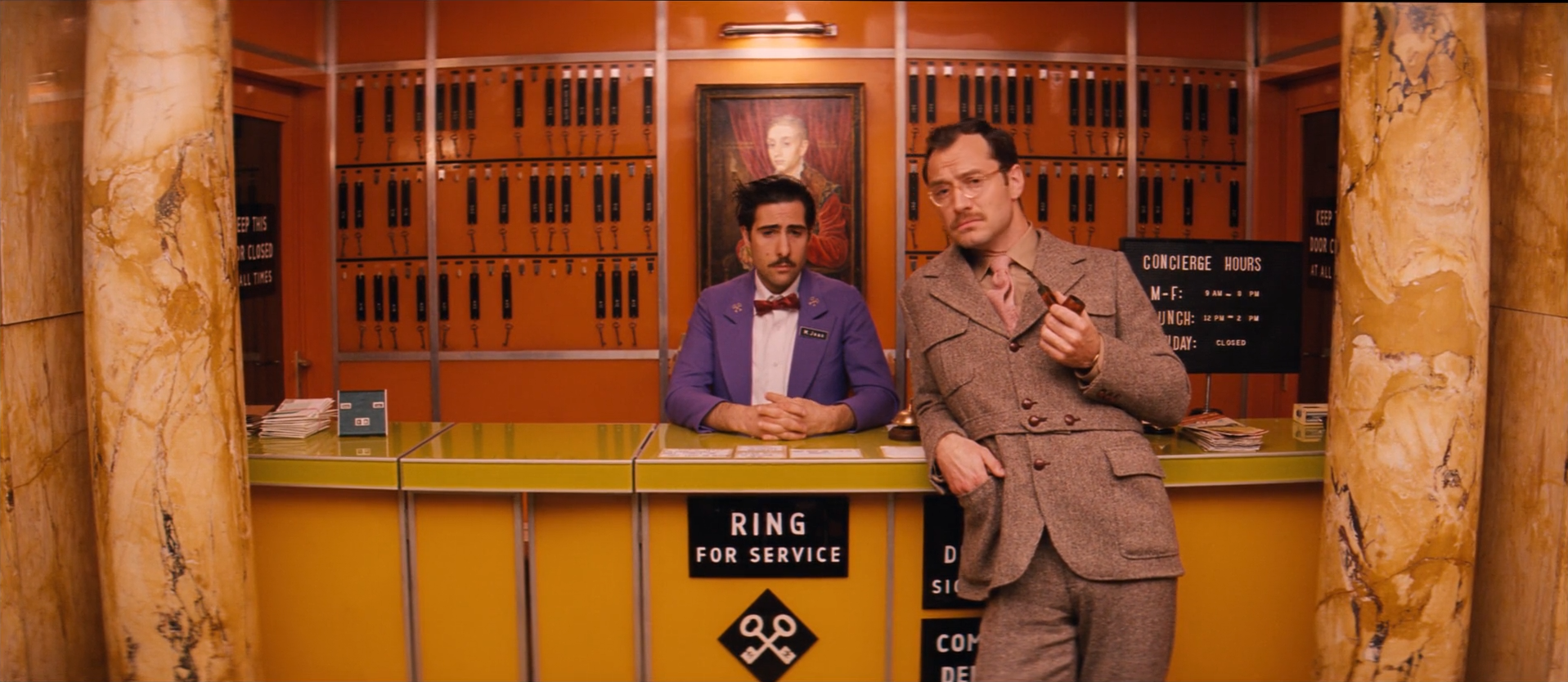 A 1968 hotel concierge desk: a man in a purple jacket and red bow tie behind a yellow-green counter reading RING FOR SERVICE, a man in a brown tweed suit holding a pipe beside him, an orange wall of key slots with a portrait painting behind, flanked by ochre marble columns.
