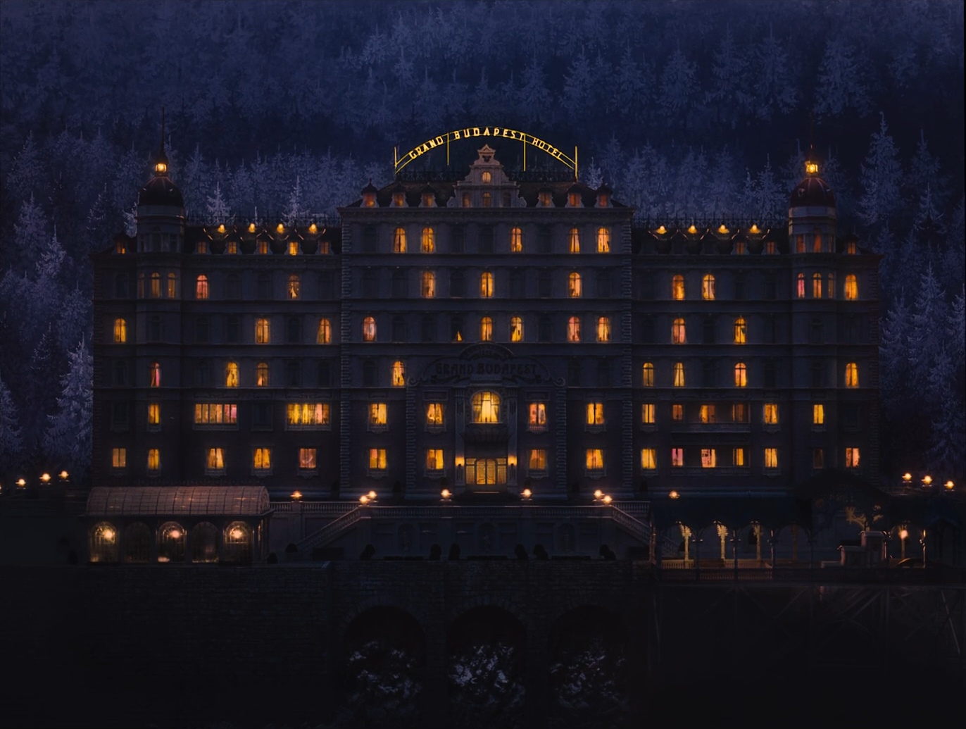 The grand hotel at night, a dark façade with rows of glowing yellow windows and a lit sign reading GRAND BUDAPEST HOTEL on the roof, against a dark blue snowy pine forest, with small lamps along the terrace.