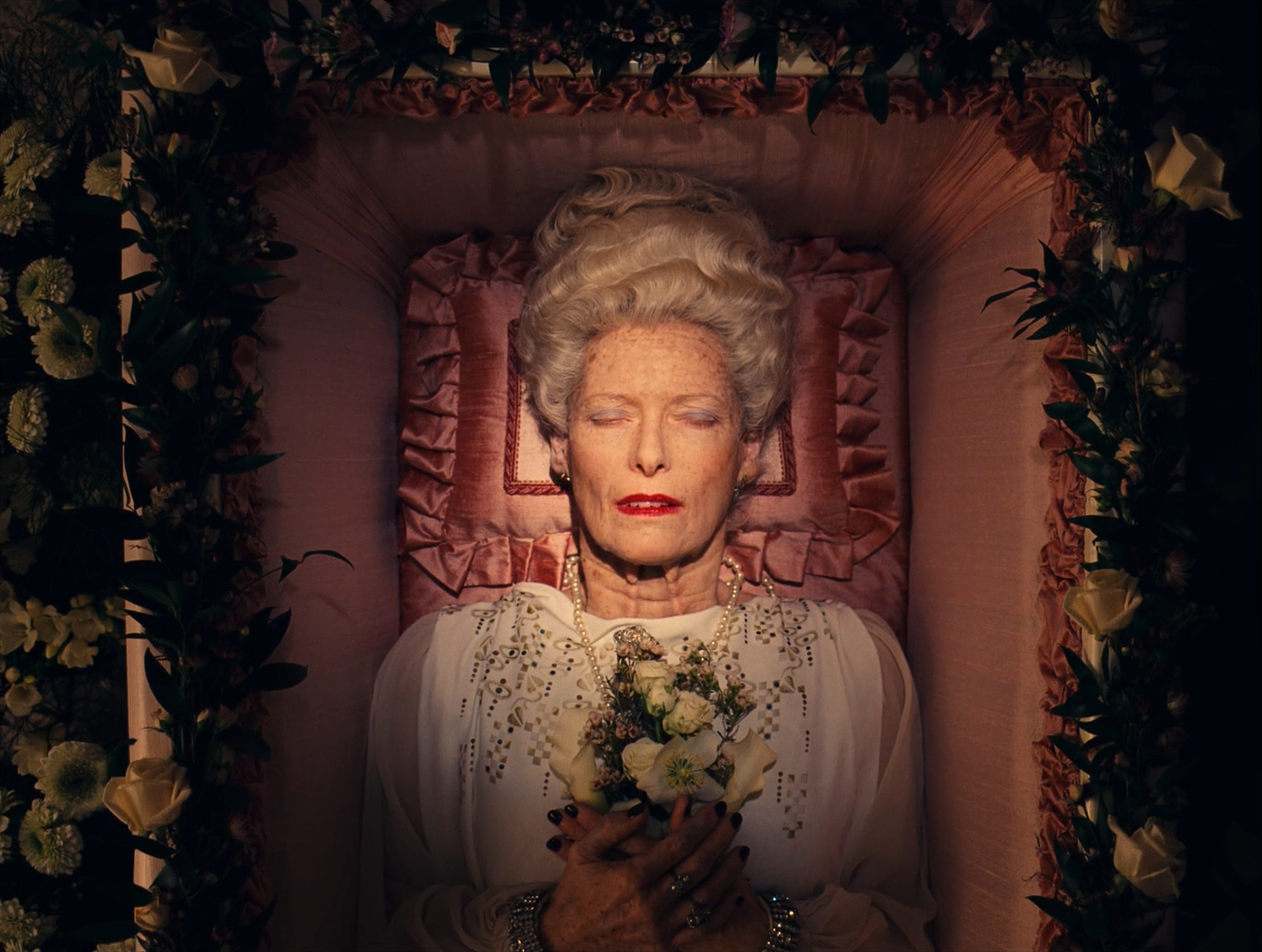 Overhead view of an elderly woman lying in an open coffin lined with pink satin, with piled white hair, red lipstick, closed eyes, a white lace dress and a pearl necklace, holding a small bouquet of white flowers, surrounded by dark greenery and white roses.