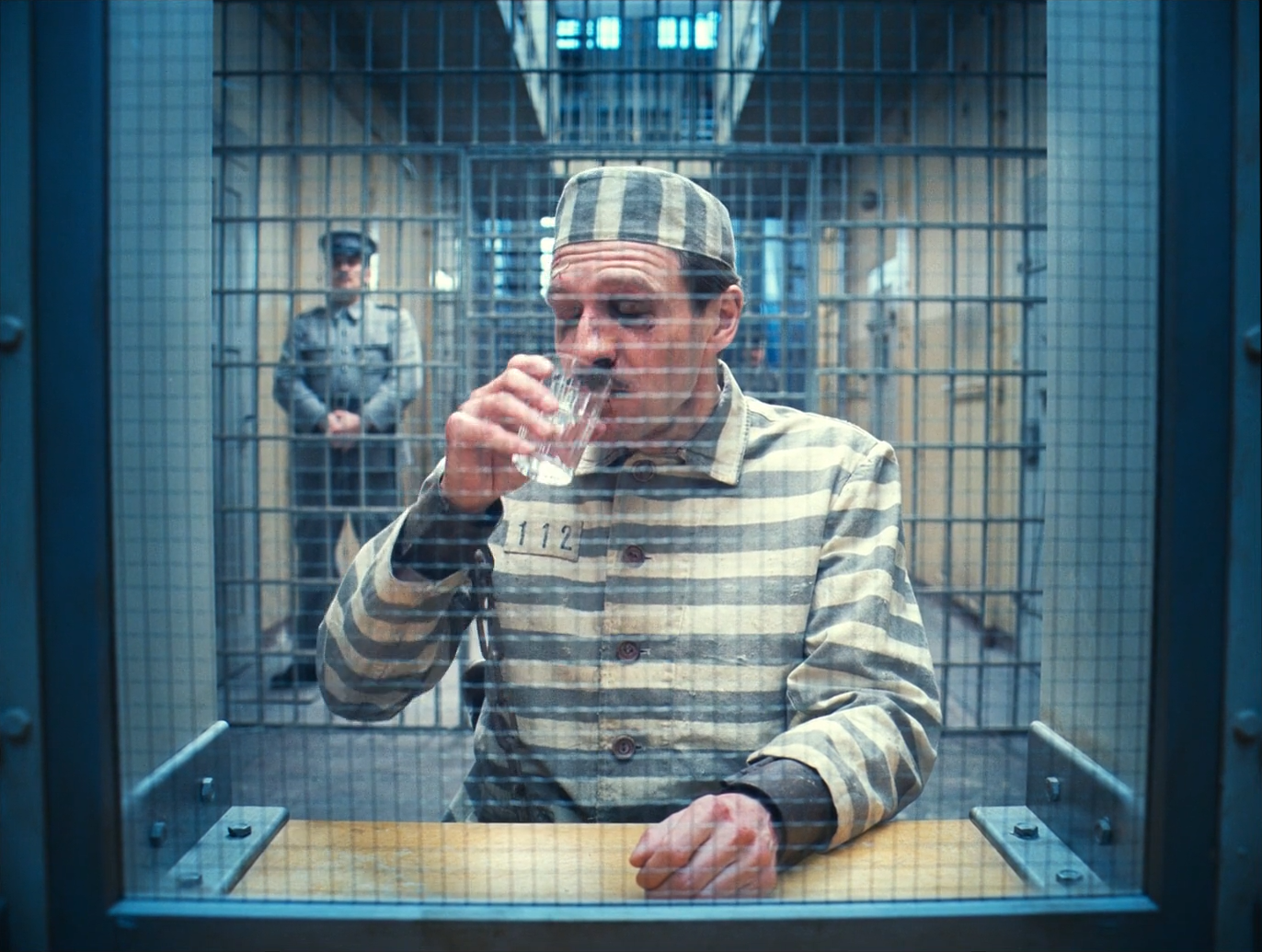 A man in a grey-and-cream striped prison uniform and cap drinks from a small glass behind a wire mesh screen with a steel frame, a uniformed guard standing behind him in front of barred cells, and a warm wooden counter at the bottom of the frame.