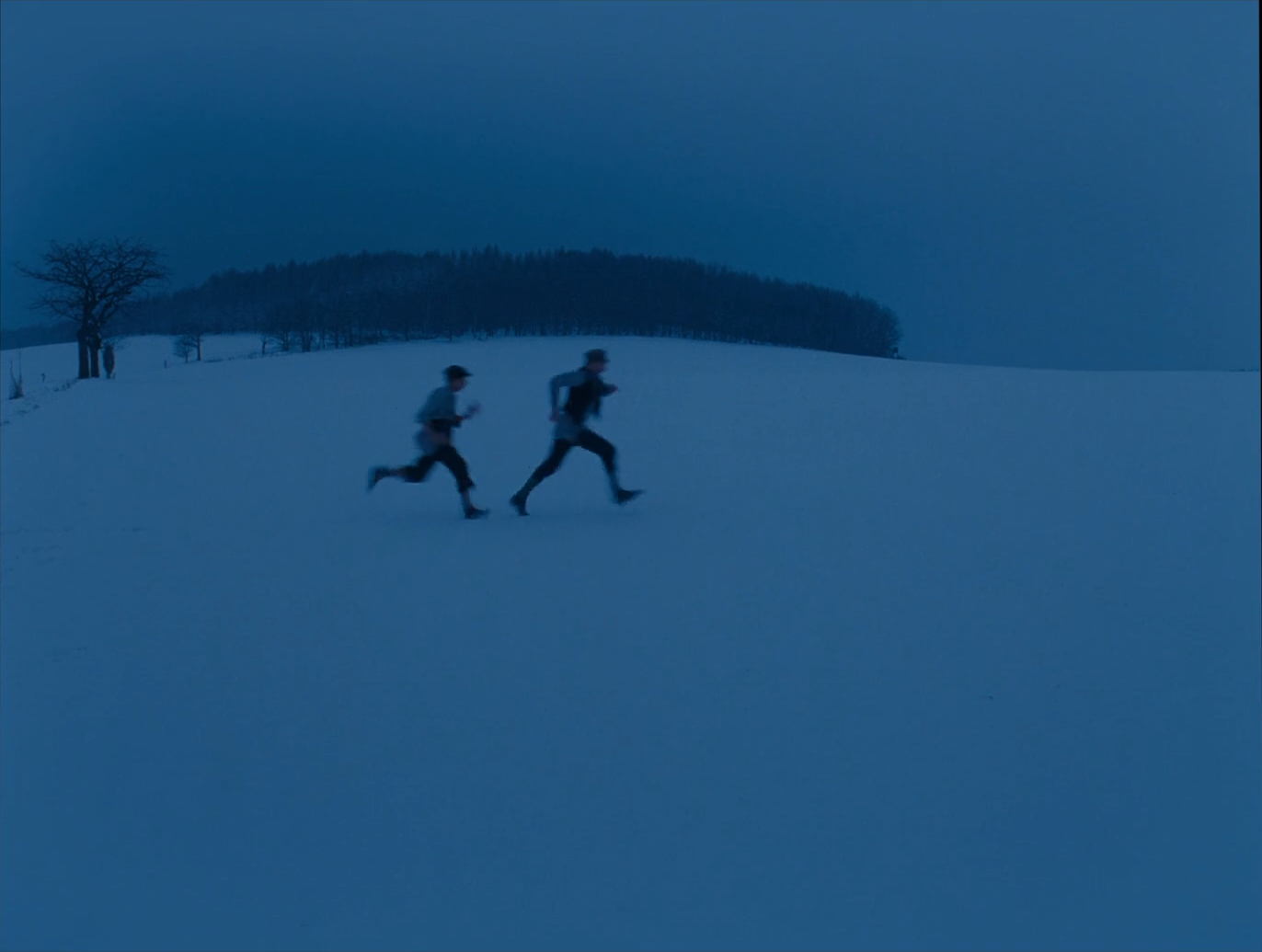 Two small figures run across a vast snowfield under a dark overcast sky, a dark line of forest along the horizon and a bare tree at the left, the whole scene in deep blue tones.
