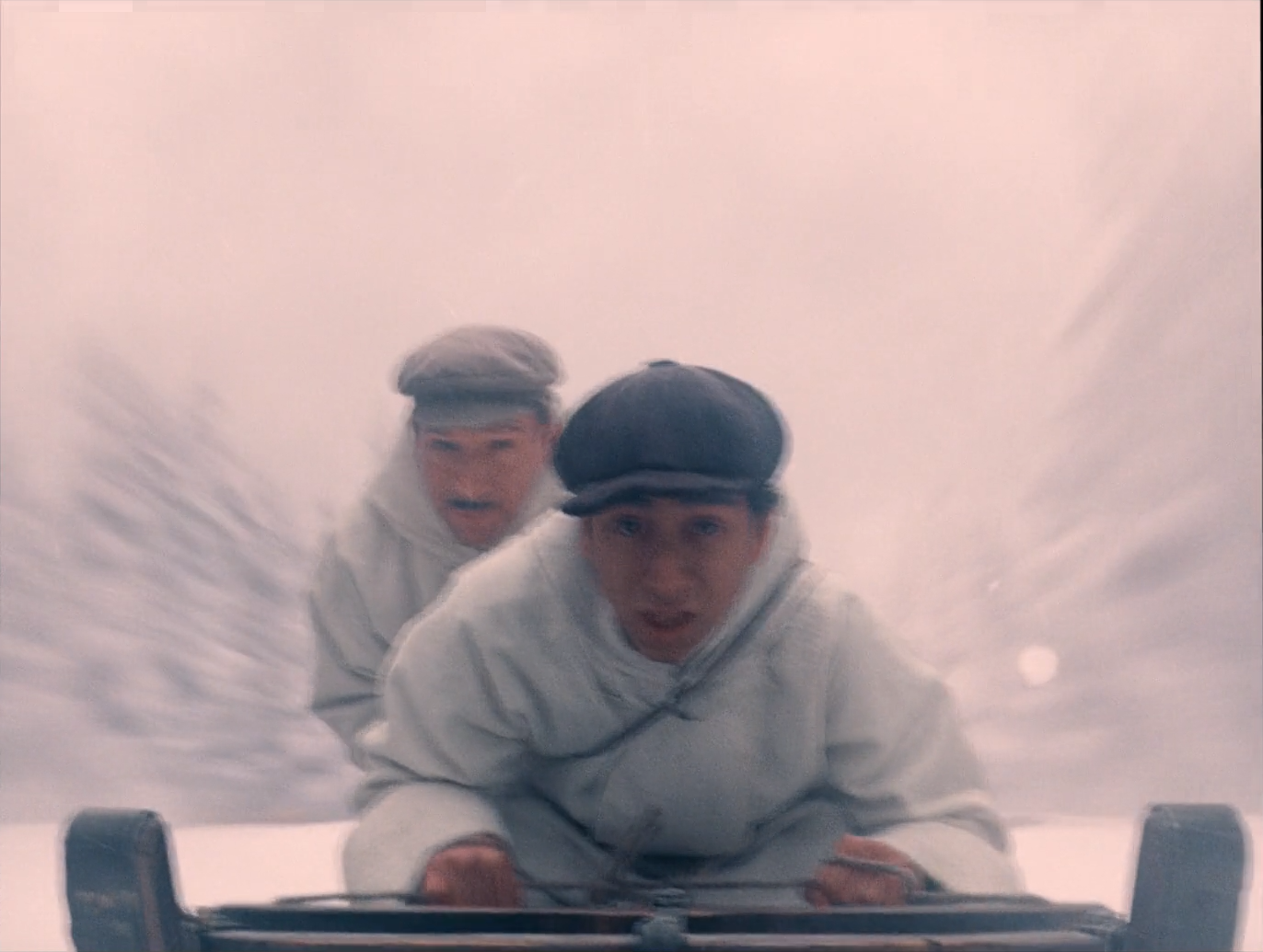 Two men in white hooded coats and dark flat caps ride a wooden sled toward the camera, faces in focus, with the snowy background streaked into pale pink-white motion blur.