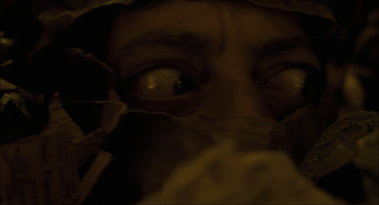 A face barely visible through dark fabric and paper, with small highlights in the eyes.