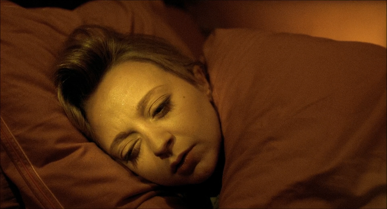A sleeping woman on rust-brown bedding, her face dimly modelled in amber shadow.