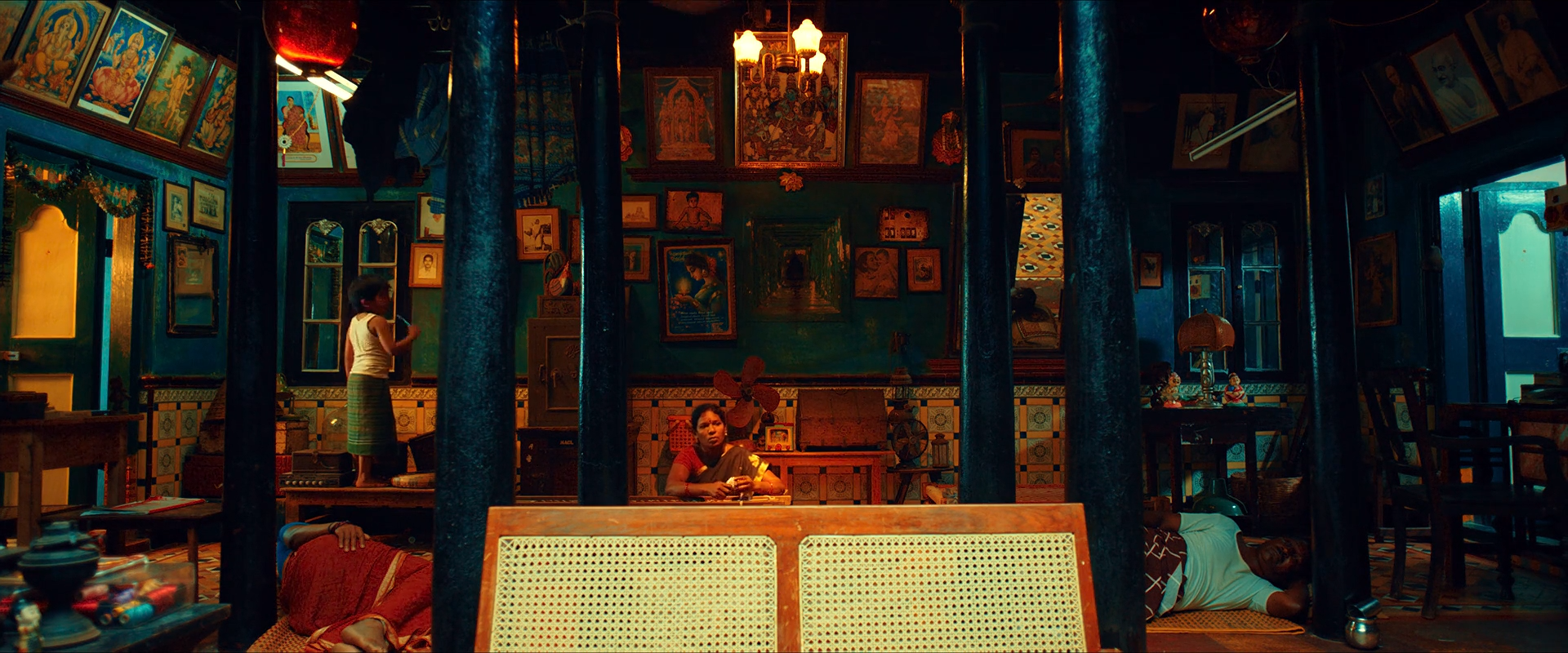 A dense devotional shop interior with teal pillars and walls, warm icons and lamps, and people arranged among dark shelves.