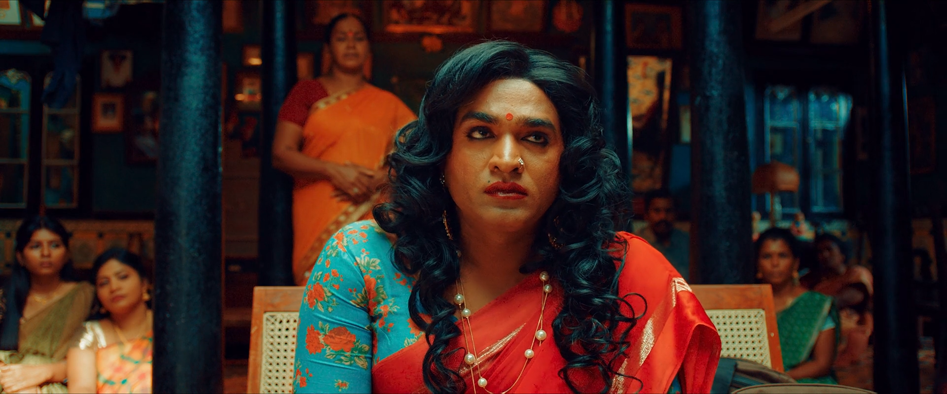 A woman with dark curls, a teal floral blouse and red sari stands in a deep teal room, her face held warm at the centre.
