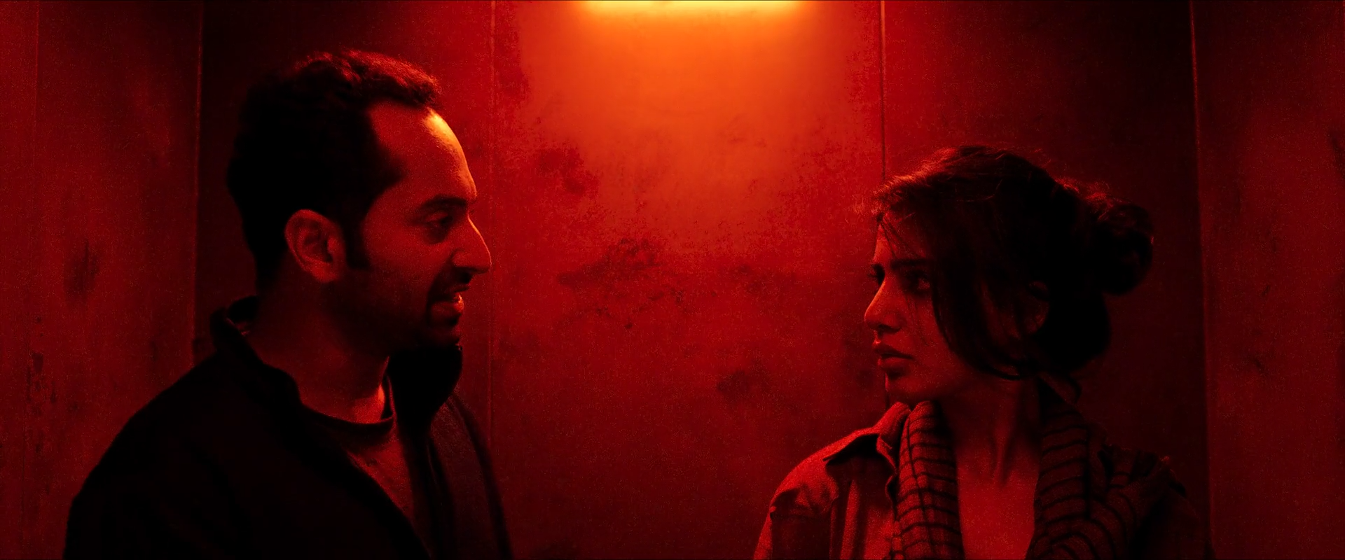 Two dark profiles face each other beneath a saturated red practical light, with a red wall and black clothing filling the frame.