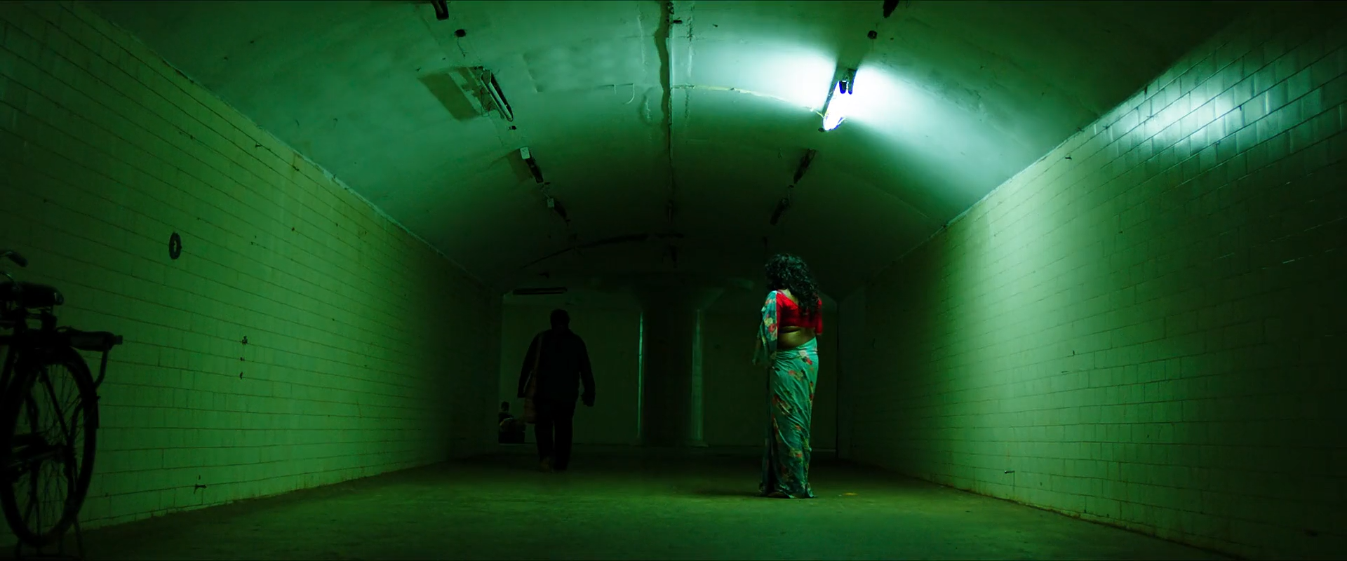 A figure in a red and teal sari walks through a long tunnel flooded by green fluorescent light.