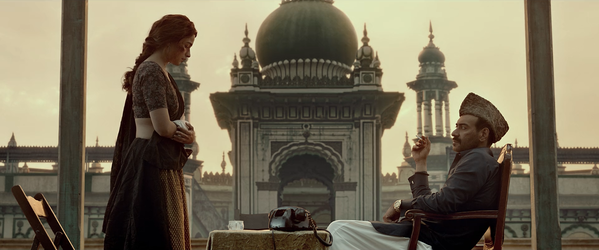 A woman and seated man stand before a monumental mosque and amber sky, with the architecture softened by haze and distance.