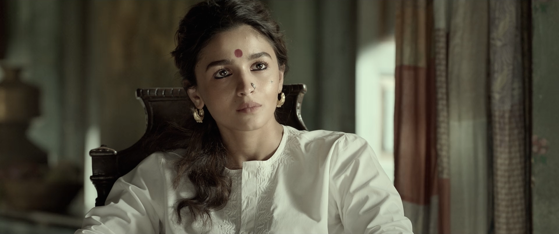 A woman in cream-white cotton sits on a dark carved chair in a muted olive room, her skin warm and softly textured.